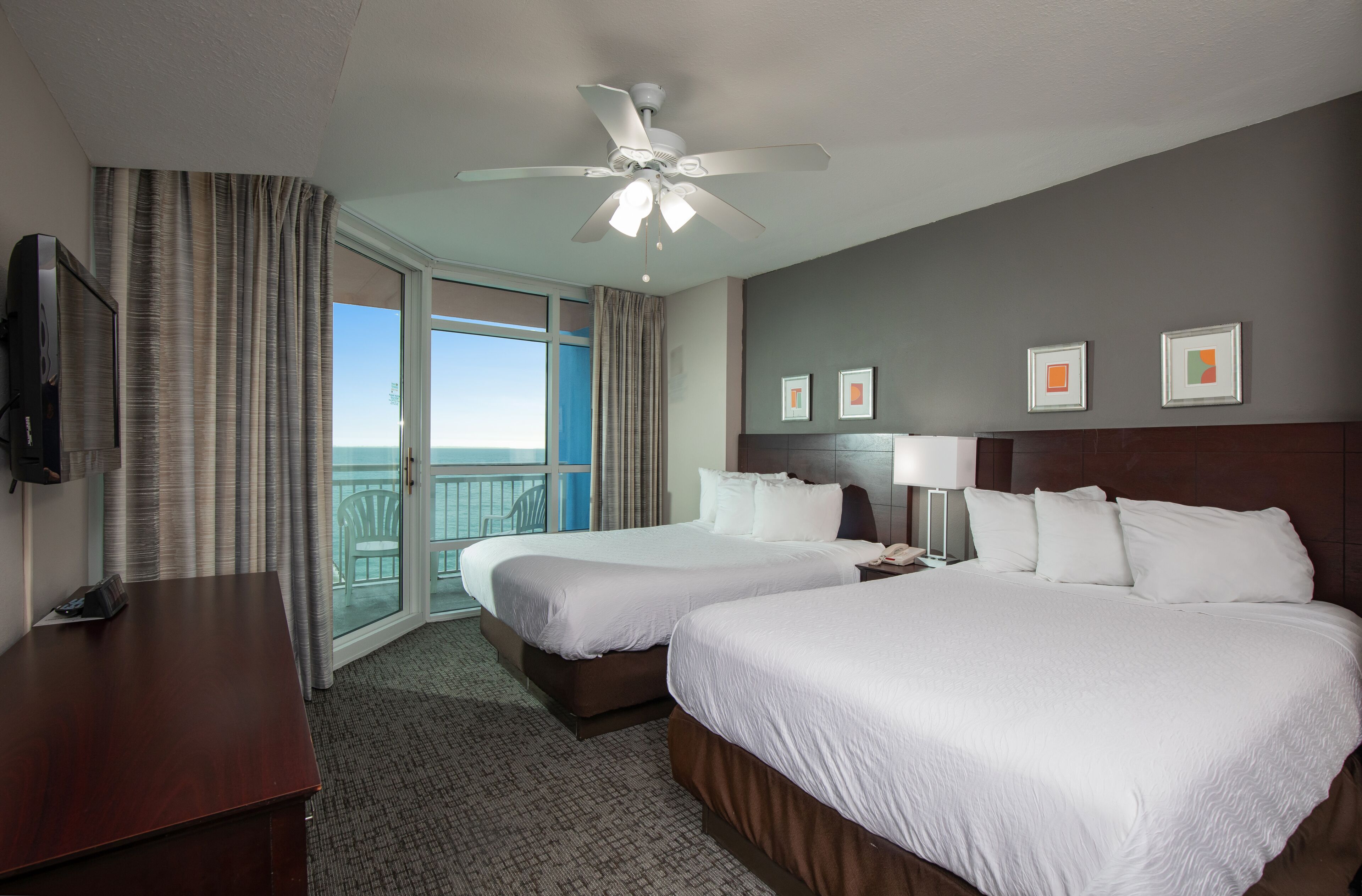 A hotel room with two beds, a desk, a TV, a ceiling fan, and a balcony with a view.