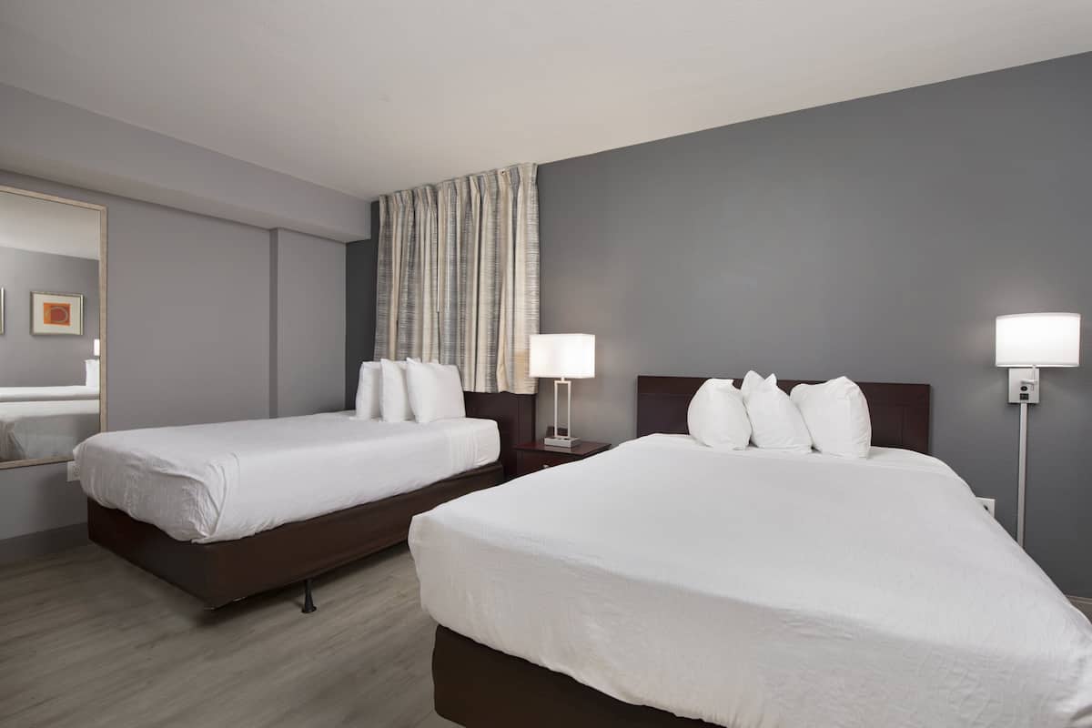 A hotel room with two single beds, a large mirror, a nightstand with a lamp, and a wall-mounted light fixture.