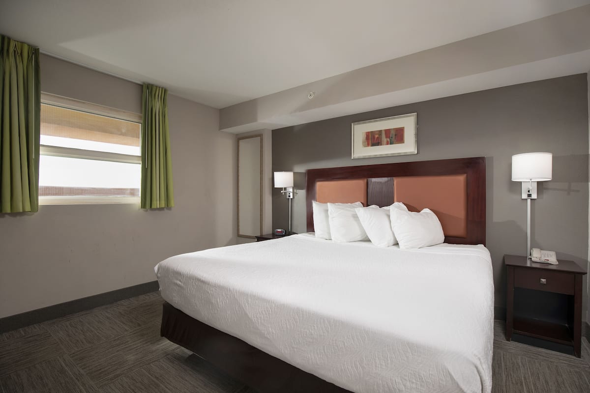 A hotel room with a large bed, two bedside tables with lamps, a window with curtains, and a framed picture on the wall.
