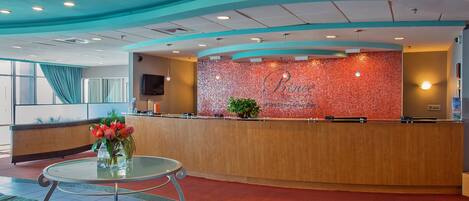 A hotel reception area with a curved reception desk, a television mounted on the wall, and a potted plant on the counter.