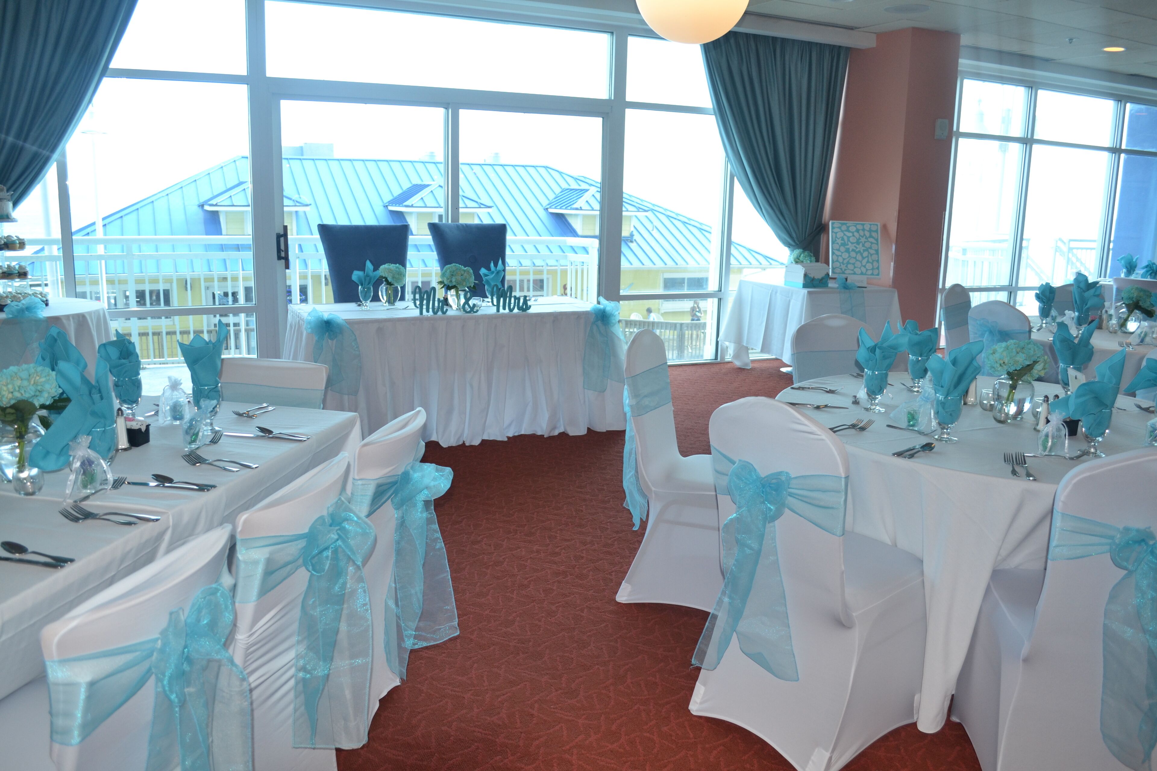 A banquet hall with white tables and chairs, each adorned with blue sashes. Large windows offer a view of a building with a blue roof.