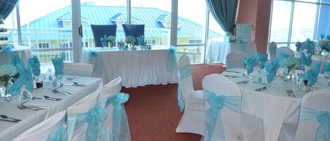 A banquet hall with white tables and chairs, each adorned with blue sashes. Large windows offer a view of a building with a blue roof.