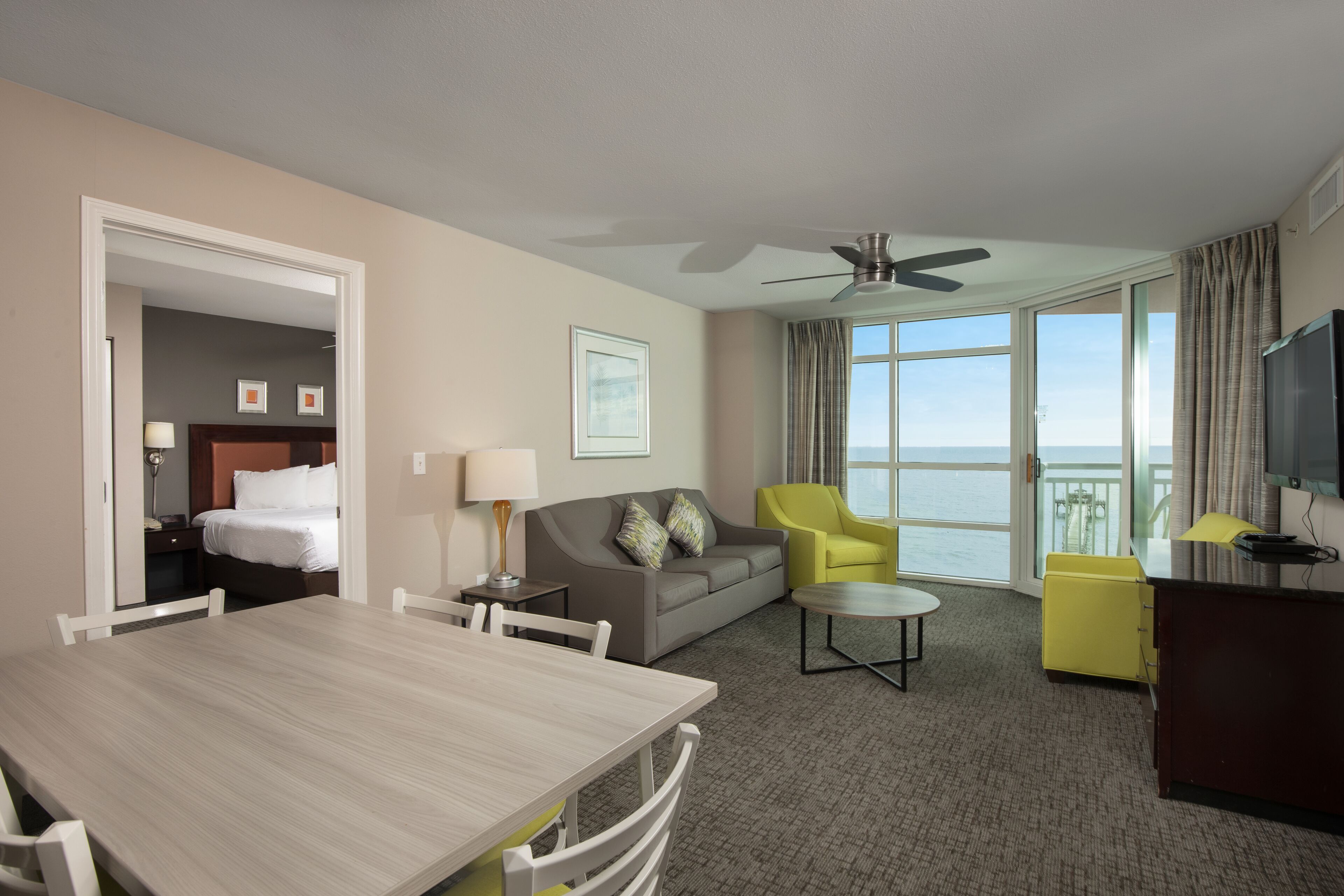 A hotel room with a dining table, a sofa, a yellow armchair, a TV, and a balcony with a view of the sea.