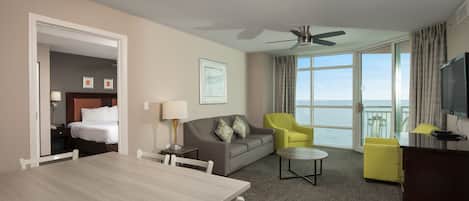 A hotel room with a dining table, a sofa, a yellow armchair, a TV, and a balcony with a view of the sea.