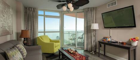 A modern living room with a sofa, yellow armchairs, a coffee table with snacks, a TV, and a balcony with a view of the ocean.