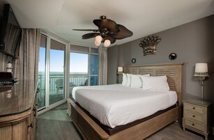 A hotel room with a large bed, a ceiling fan, a TV, a nightstand, and a view of the ocean.
