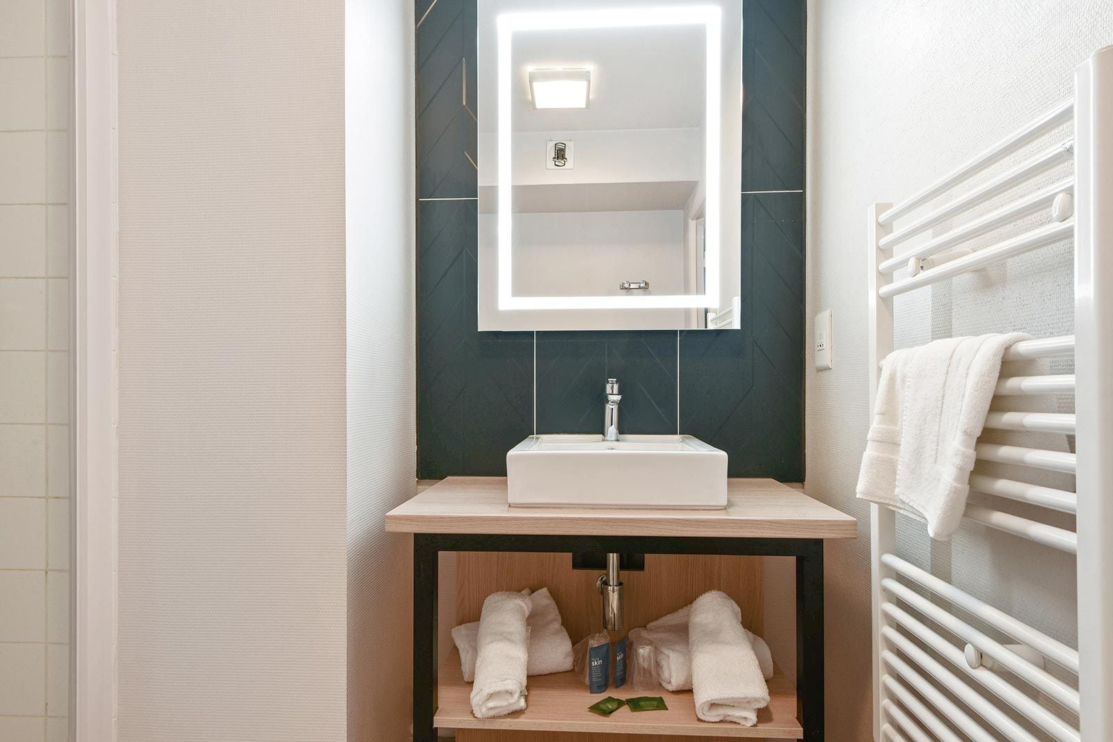 A bathroom with a white sink, a mirror with built-in lighting, a towel rack, and a shelf with towels and toiletries.