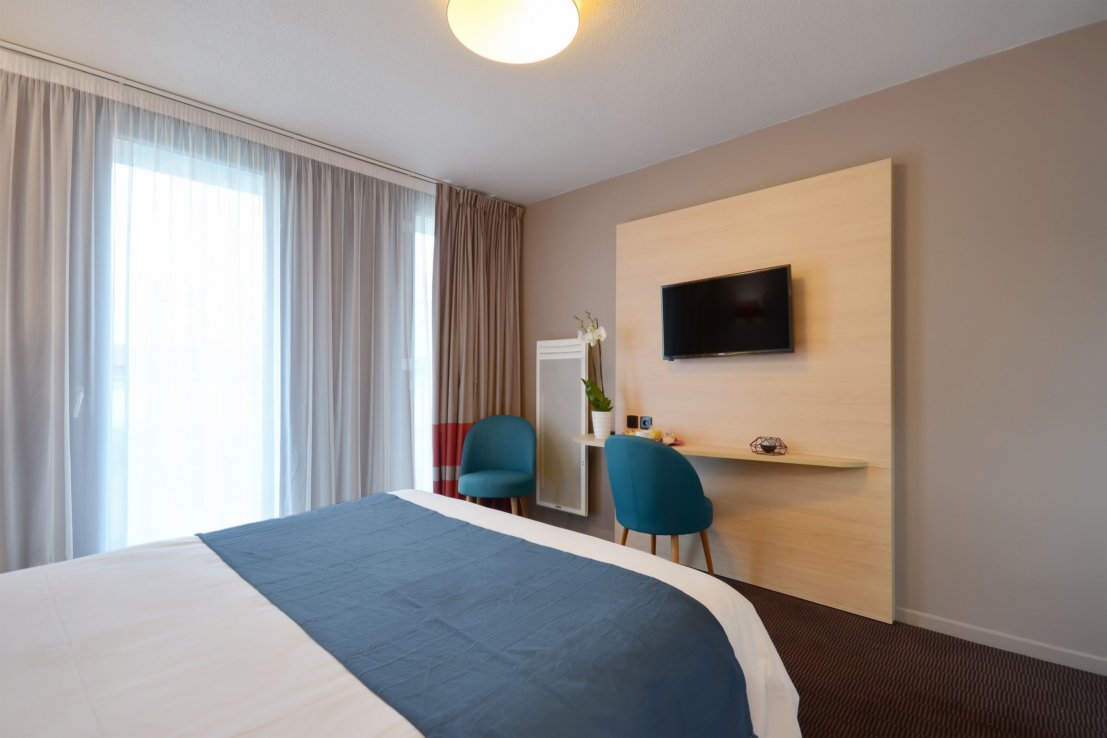 A hotel room with a large bed, two blue chairs, a TV mounted on the wall, and a desk with a plant.