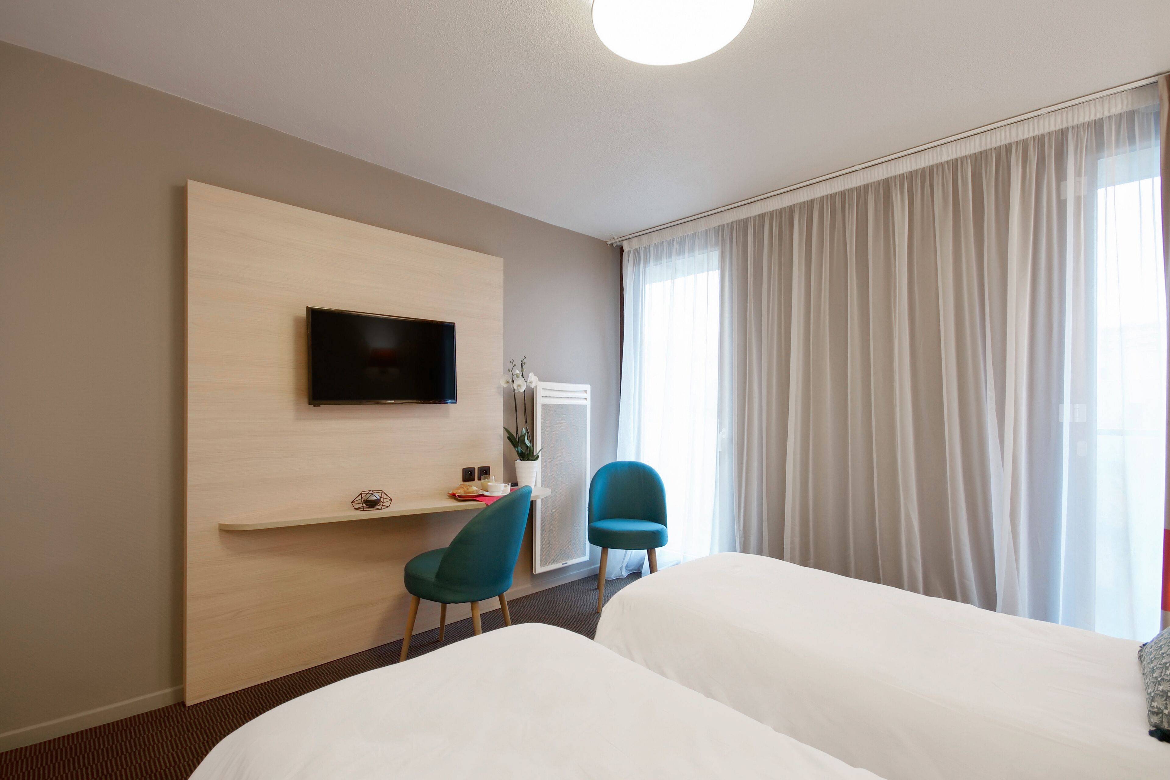 A hotel room with a bed, a desk with a chair, a TV mounted on the wall, and a large window with curtains.