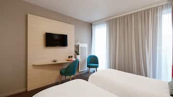 A hotel room with a bed, a desk with a chair, a TV mounted on the wall, and a large window with curtains.
