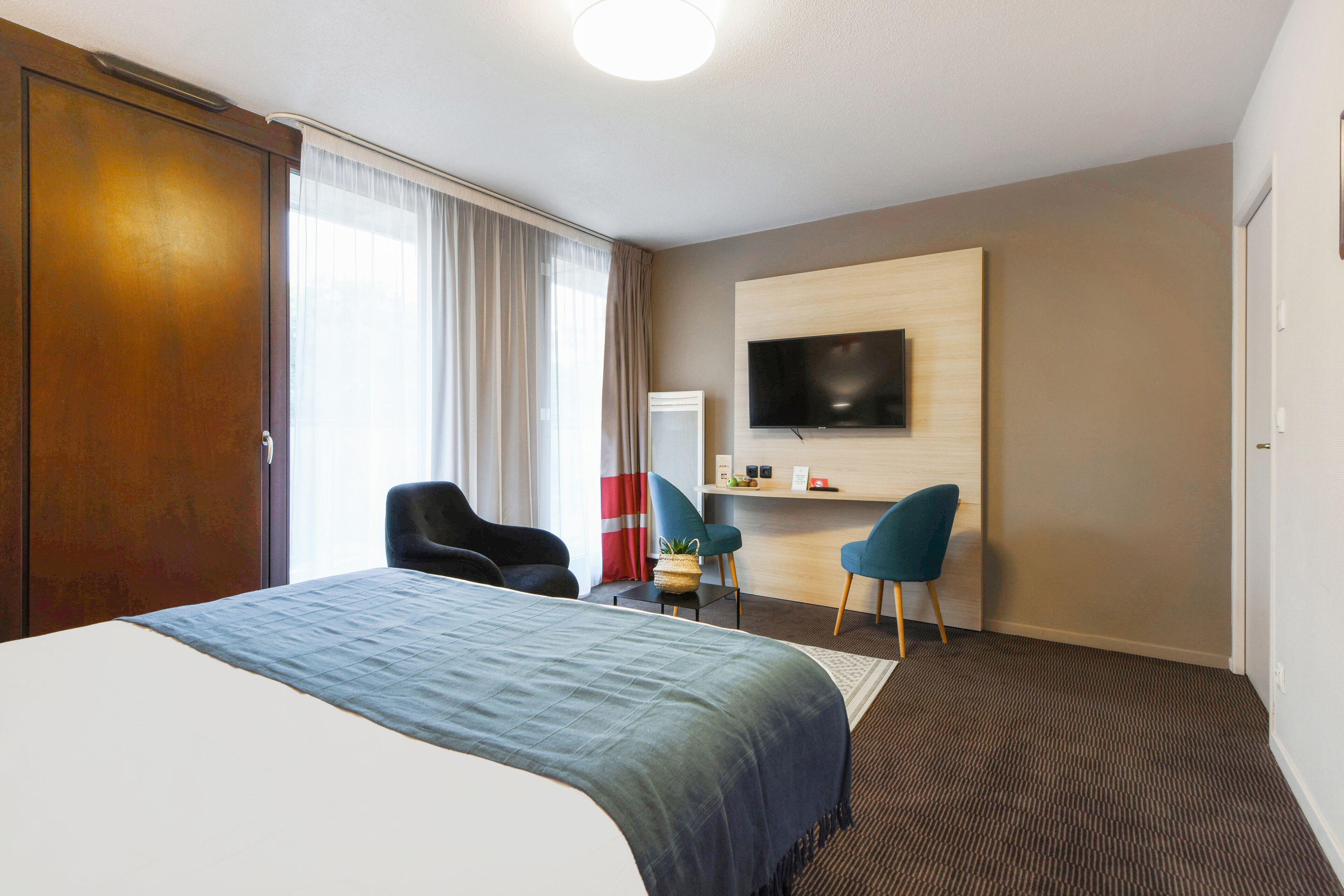 Une chambre d’hôtel avec un grand lit, un bureau avec une chaise, un téléviseur fixé au mur et une fenêtre avec des rideaux.