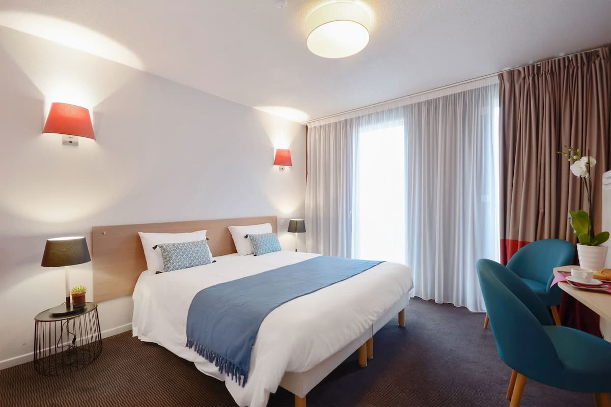 Une chambre d’hôtel avec un lit, deux lampes de chevet, un bureau avec une chaise, une plante en pot et une fenêtre avec des rideaux.