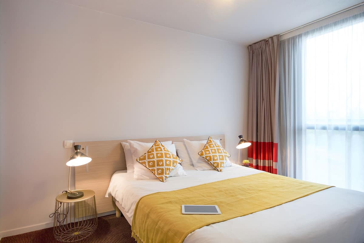 Une chambre d’hôtel avec un lit, deux lampes de chevet, une table de chevet, une tablette et une fenêtre avec des rideaux.