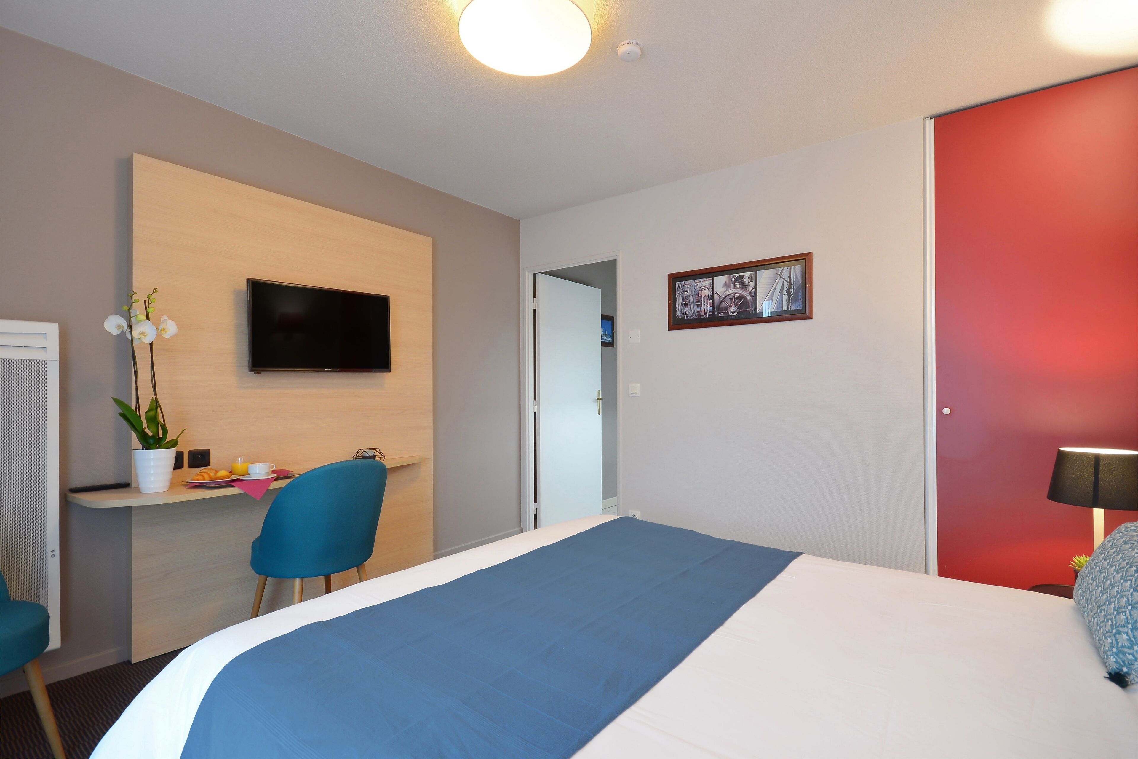 A hotel room with a bed, a desk with a chair, a television, and a red door.