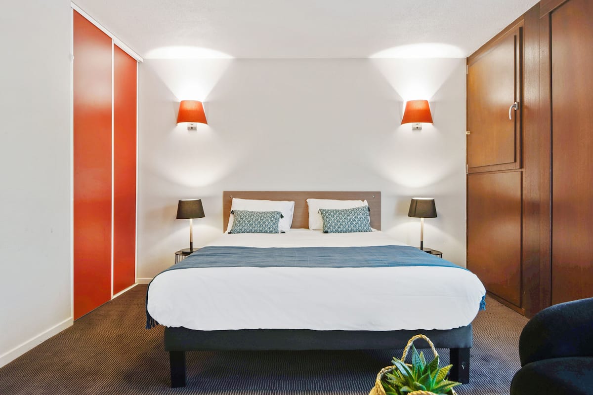 Une chambre d’hôtel avec un lit, deux lampes de chevet, une tête de lit, une armoire et une plante en pot.