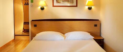 A hotel room with a wooden headboard, two white pillows, a bed, a nightstand, two wall-mounted lamps, and a framed picture on the wall.