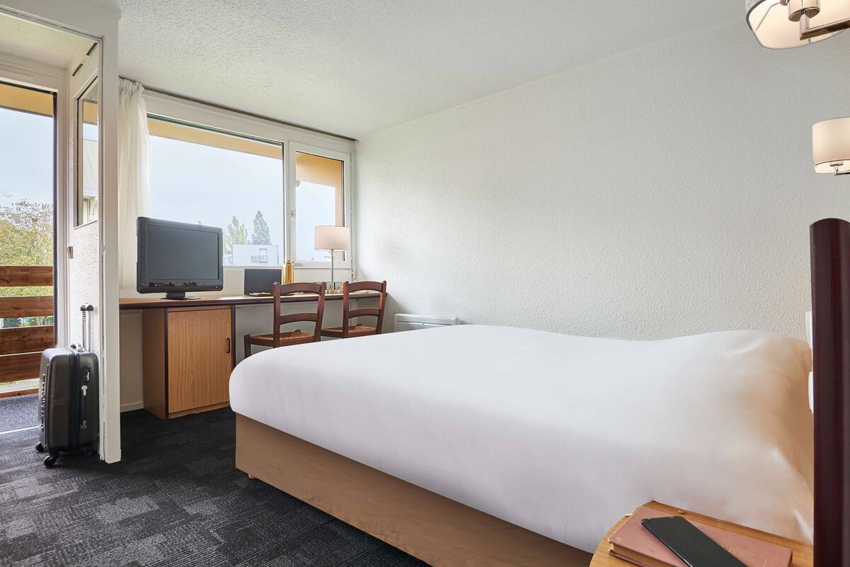 A hotel room with a bed, a desk with a computer, a chair, a lamp, and a suitcase.