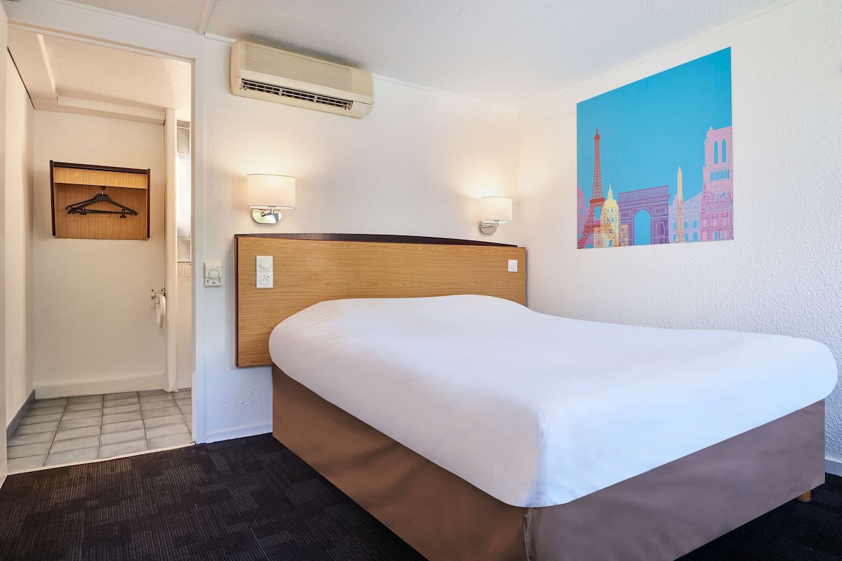 A hotel room with a single bed, a wooden headboard, two wall-mounted lamps, an air conditioning unit, a framed picture of Paris landmarks, and a door leading to another room.