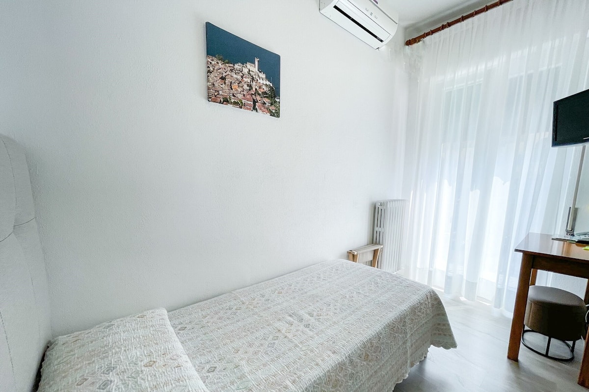 A single bed with a white bedspread, a small bedside table, a chair, a radiator, a TV, and a window with sheer curtains.