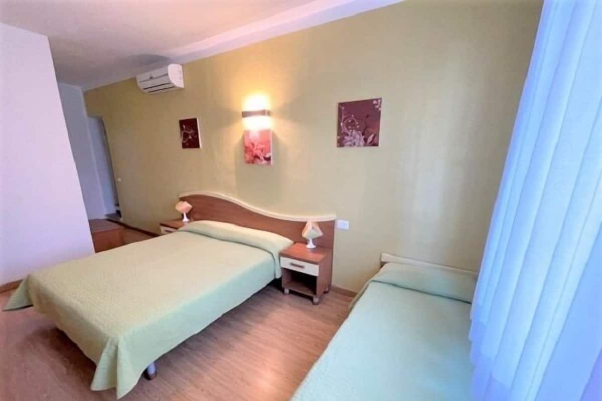 A hotel room with two single beds, a nightstand with a lamp, a wall-mounted air conditioner, and two wall art pieces.