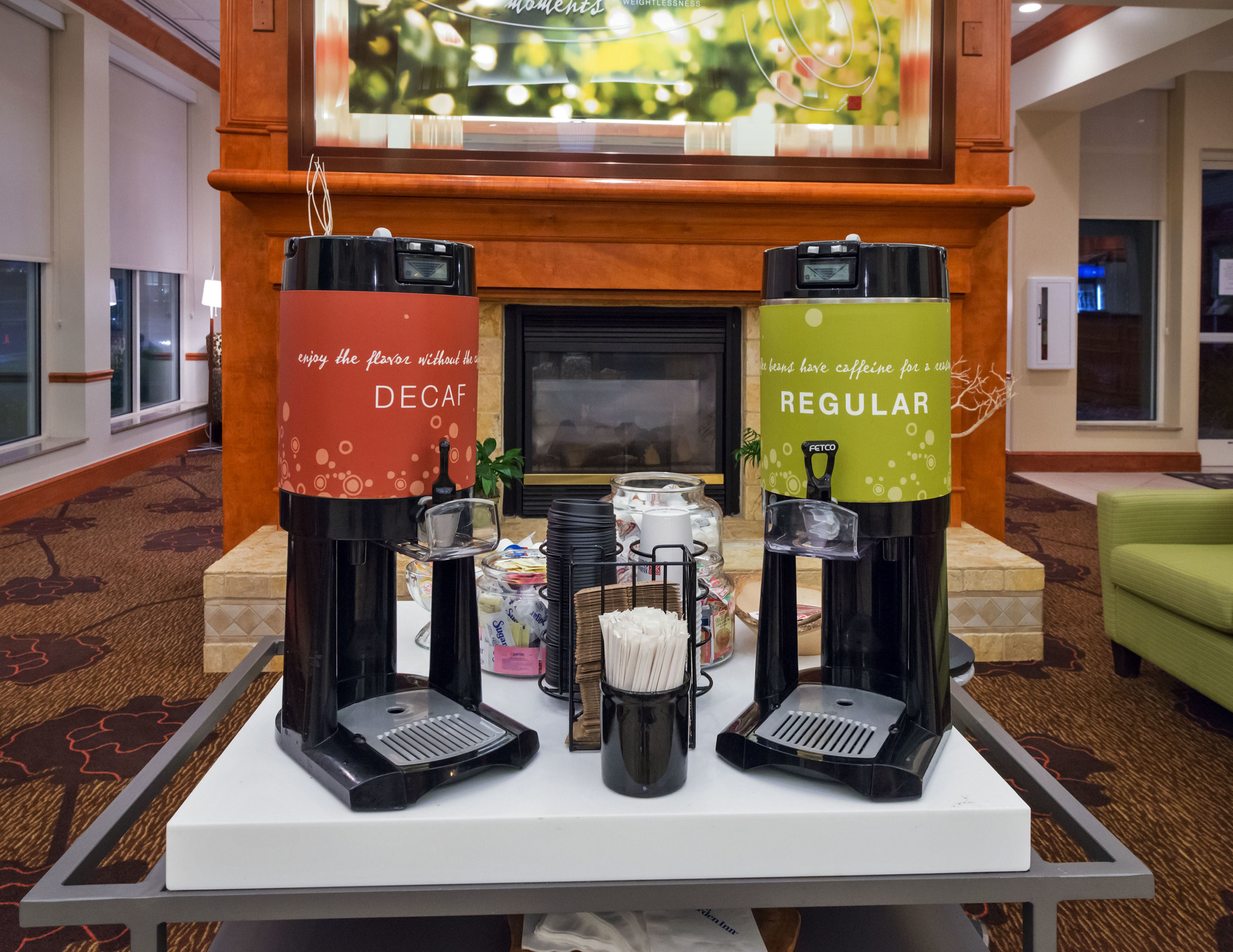 En kaffestation med to maskiner til skattefri og almindelig kaffe, en kaffekande samt forskellige kaffekapsler og redskaber på et bord i en hotellobby.