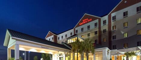 A Hilton Garden Inn with a well-lit entrance, a covered parking area, and a landscaped outdoor seating area.