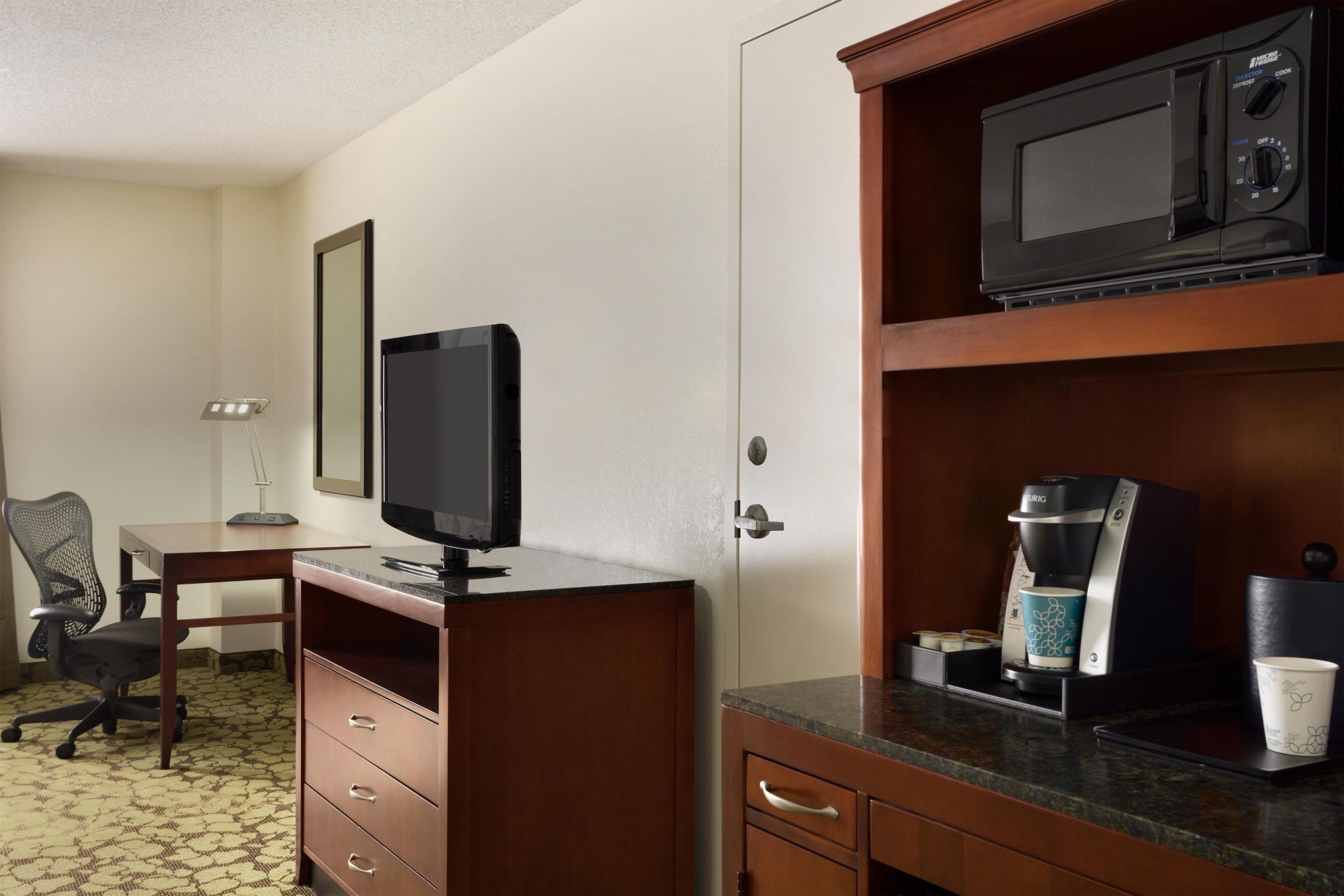 A hotel room with a flat-screen TV, a coffee maker, a microwave, a desk, and a chair.