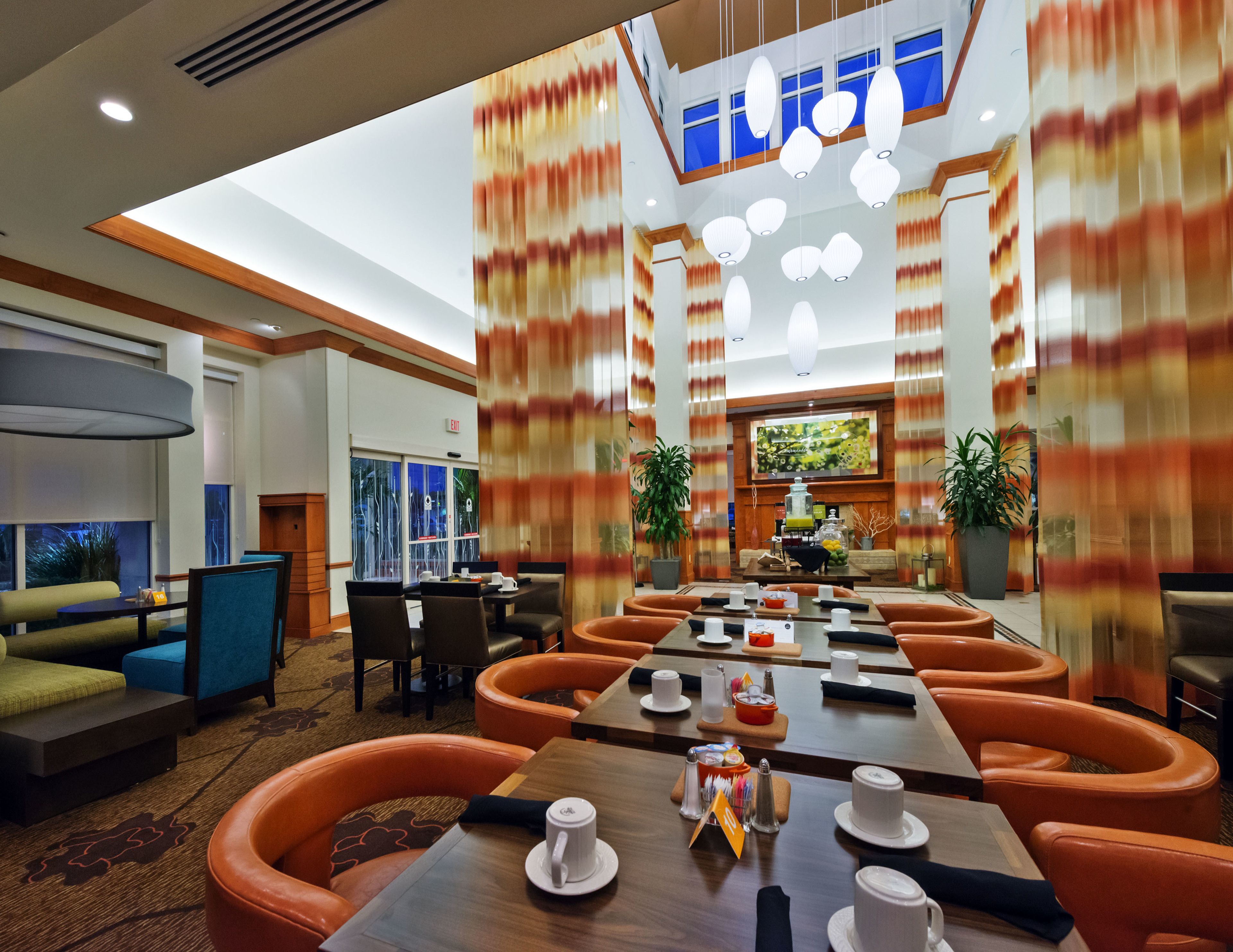Un espace de restauration d’hôtel avec plusieurs tables et des banquettes orange rembourrées, une grande fenêtre offrant une vue et un écran de télévision fixé au mur.