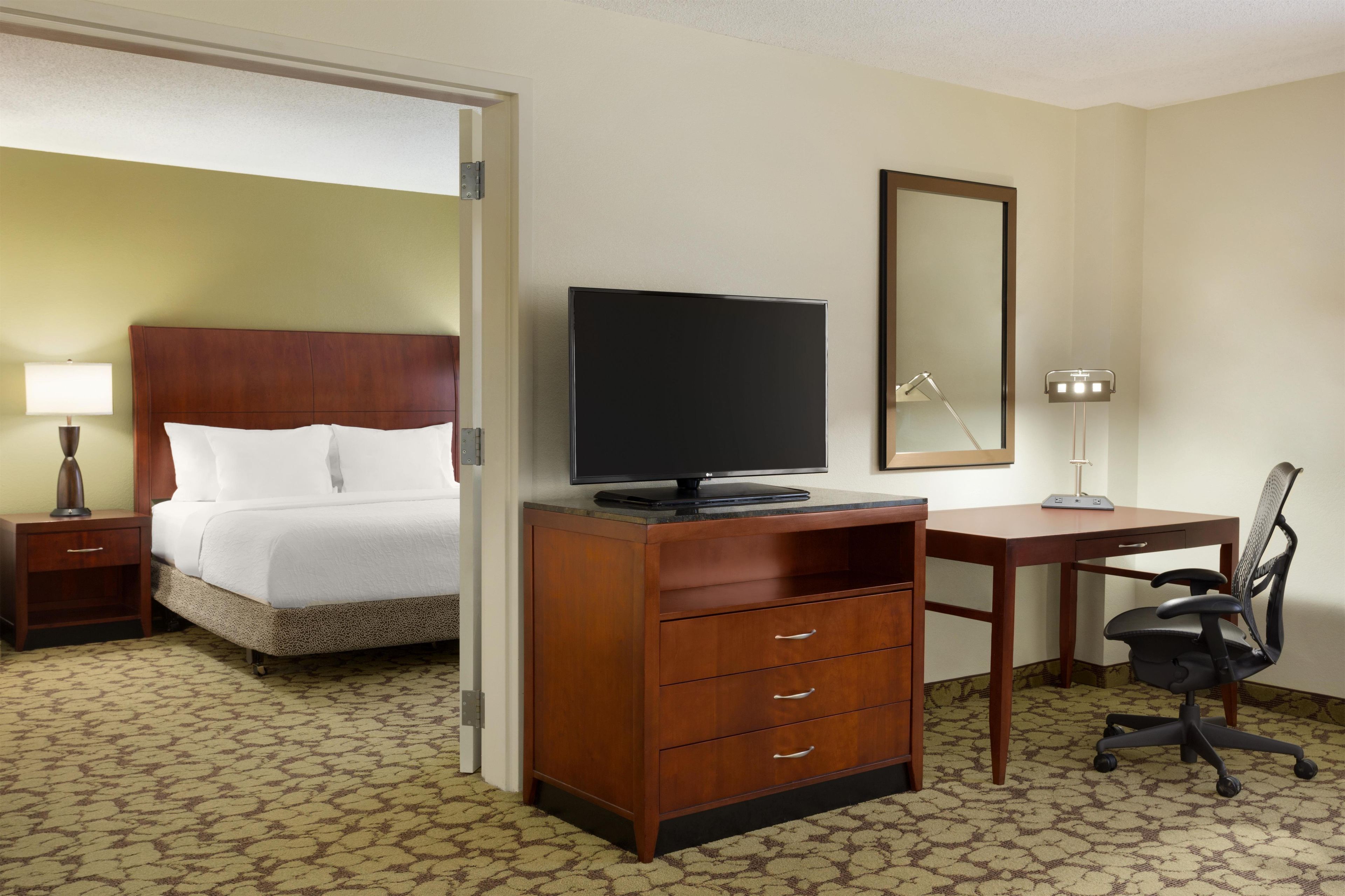 Habitación de hotel con cama, escritorio, silla, televisor y espejo.