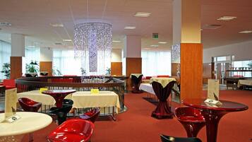 Een modern restaurant met ronde tafels en rode stoelen, met een bar in het midden en een decoratieve hanglamp.