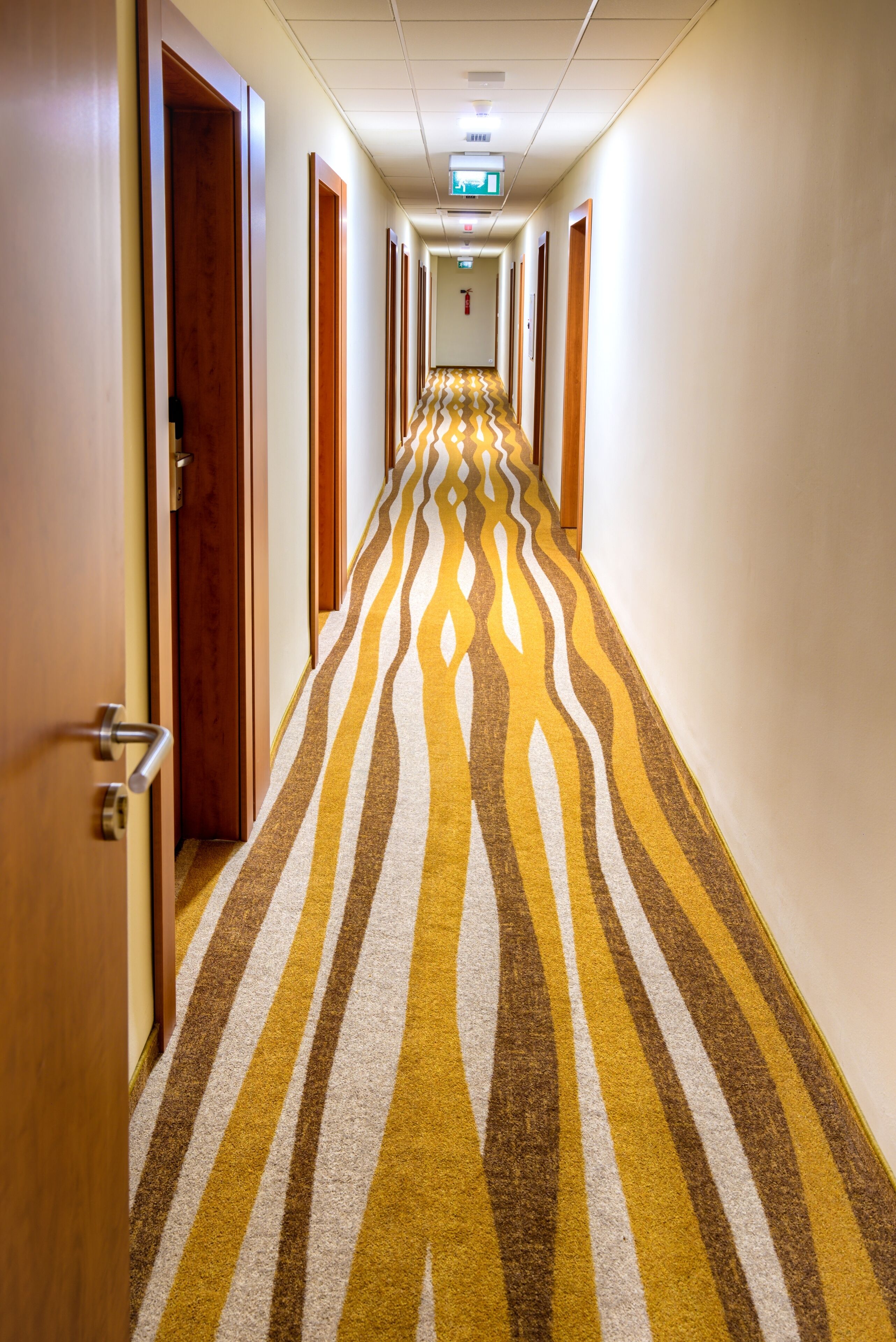 A hotel hallway with a patterned carpet, doors on both sides, and emergency exit signs on the ceiling.