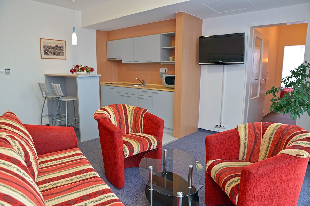 A compact living space with a kitchenette, two red striped sofas, a small round glass table, and a flat-screen TV mounted on the wall.