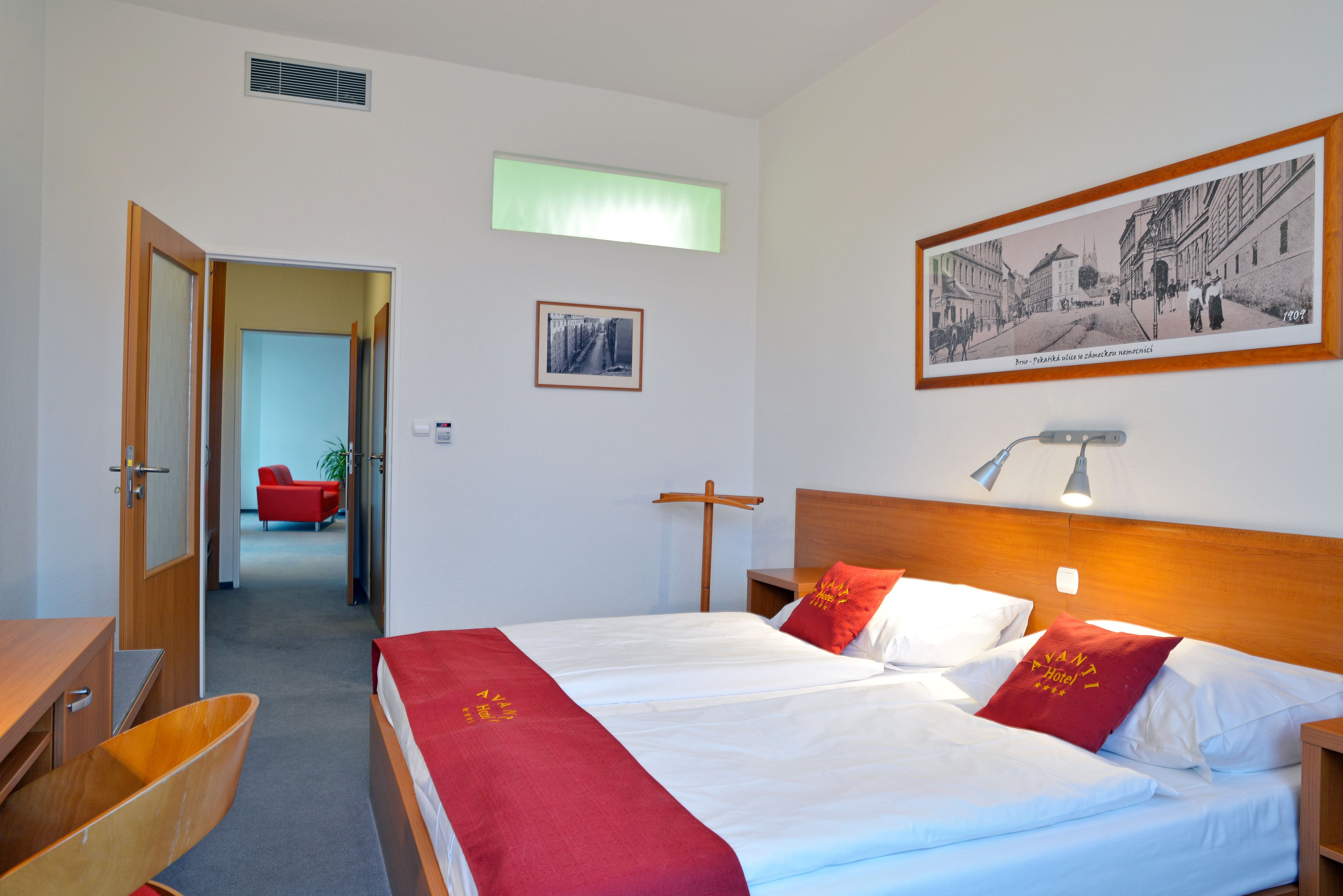 A hotel room with a large bed, a wooden headboard, a desk, and a chair. There is a window with a view of the street and a framed picture on the wall.