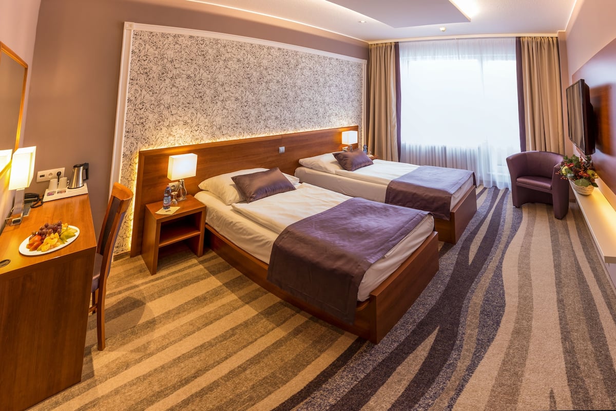 A hotel room with two beds, a desk with a lamp, a chair, a TV, and a window with curtains.