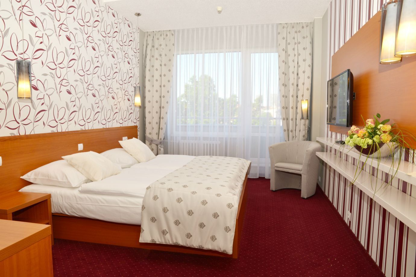 A hotel room with a large bed, a chair, a TV, and a flower vase.