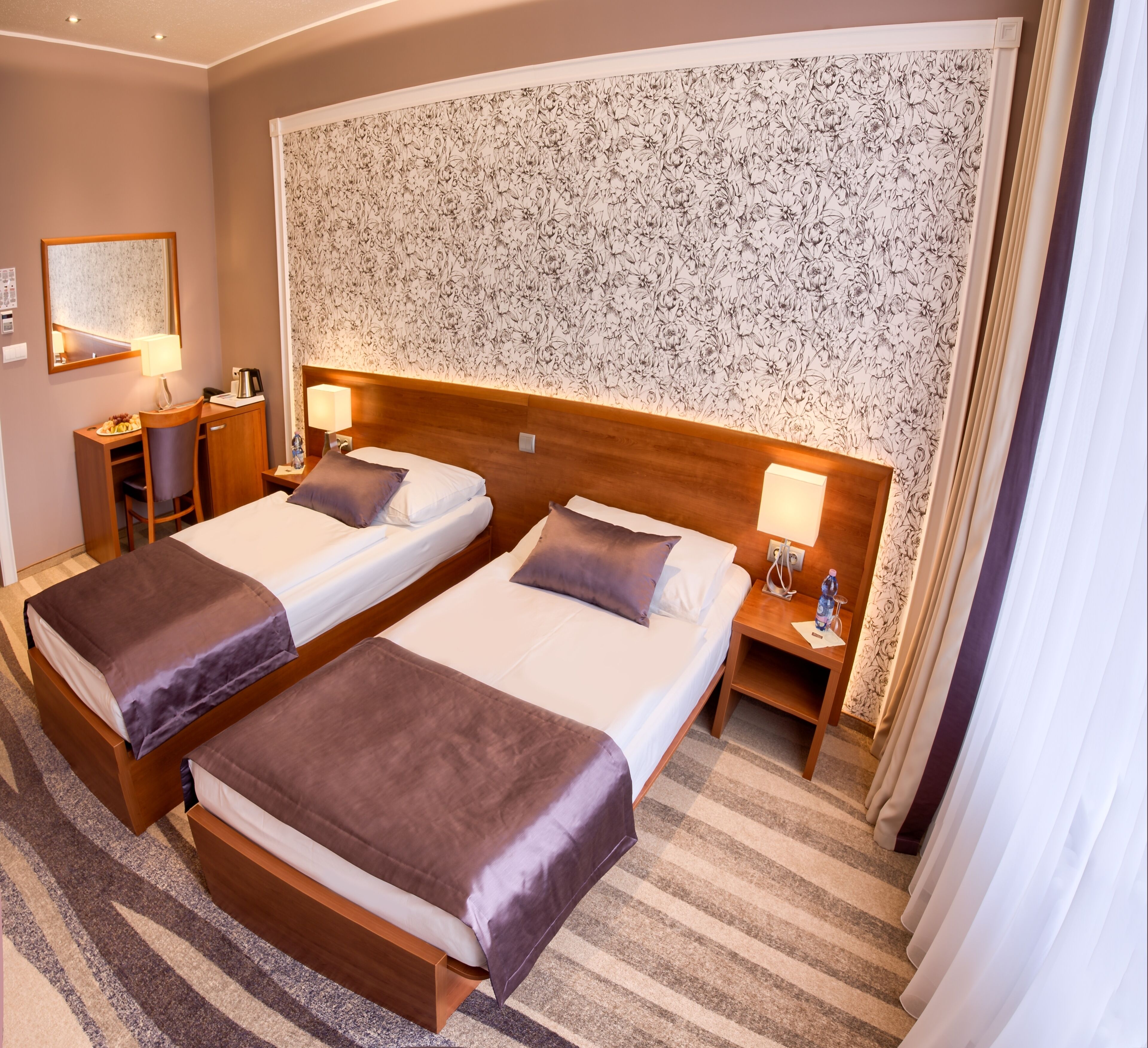 A hotel room with two beds, a desk, a chair, a mirror, and a floral wallpaper.