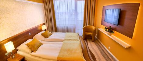 A hotel room with two beds, a TV mounted on the wall, a small table with a lamp, and a window with curtains.