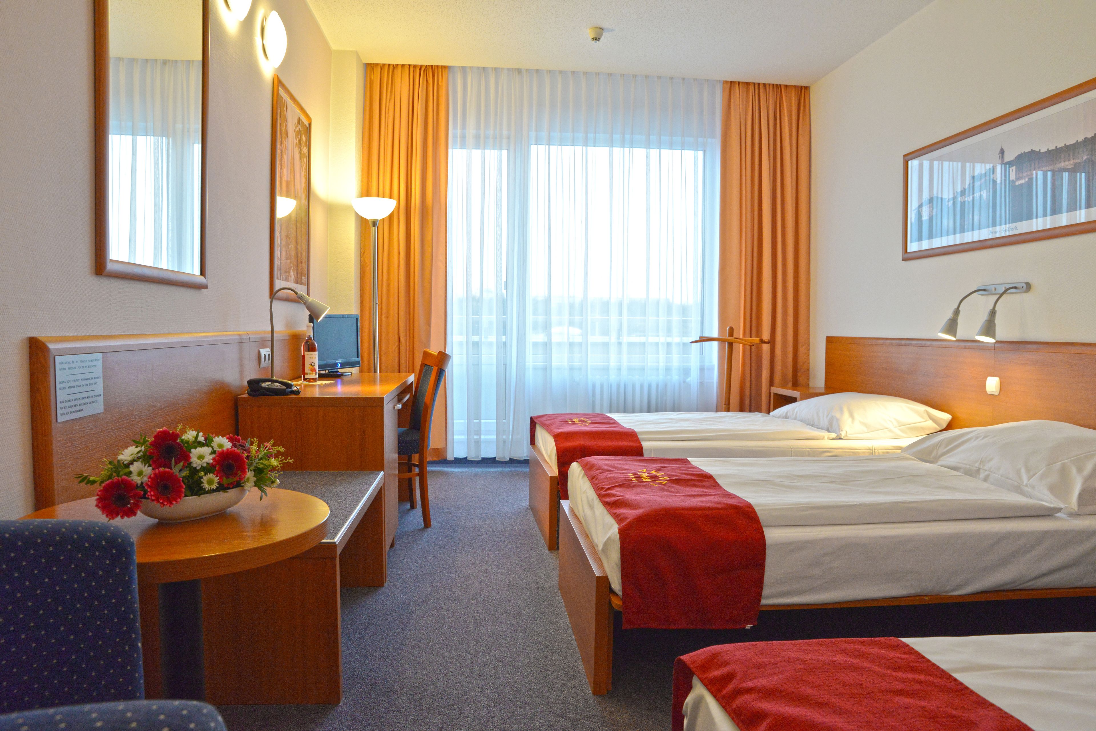 A hotel room with two beds, a desk with a chair, a small table with a flower vase, and a large window with curtains.