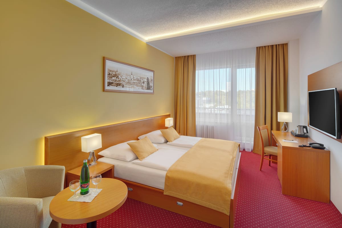 A hotel room with a double bed, a desk with a chair, a television, and a small table with a bottle and glasses.