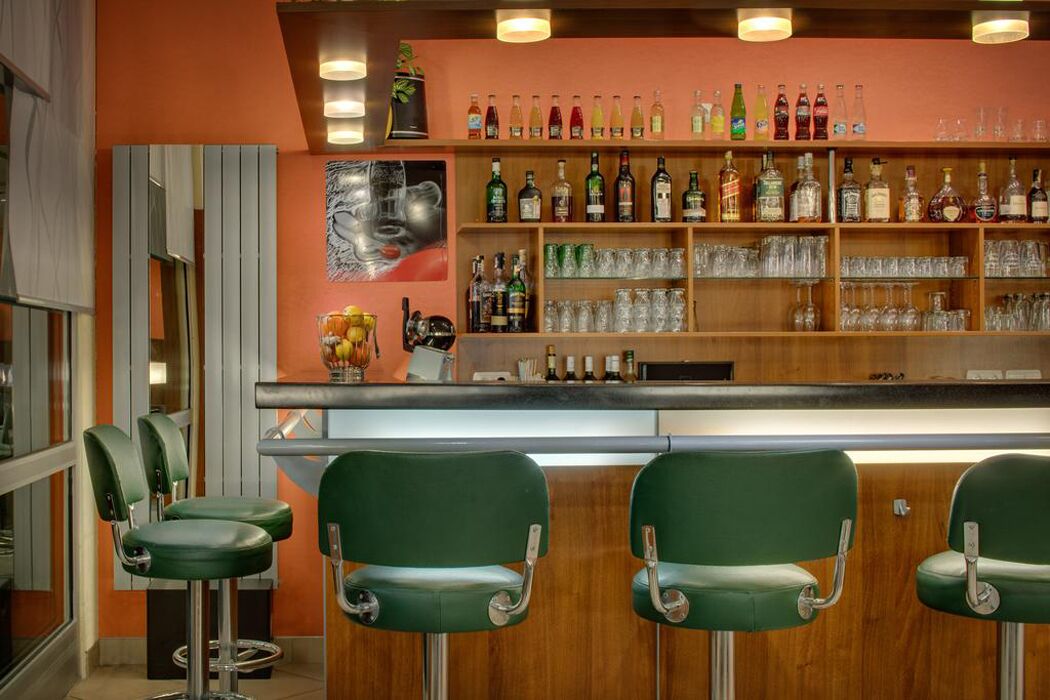 Eine Bar mit grünen Barhocker, einem Holzbodenregal mit verschiedenen Flaschen und Gläsern sowie einem an der Wand hängenden Gemälde.