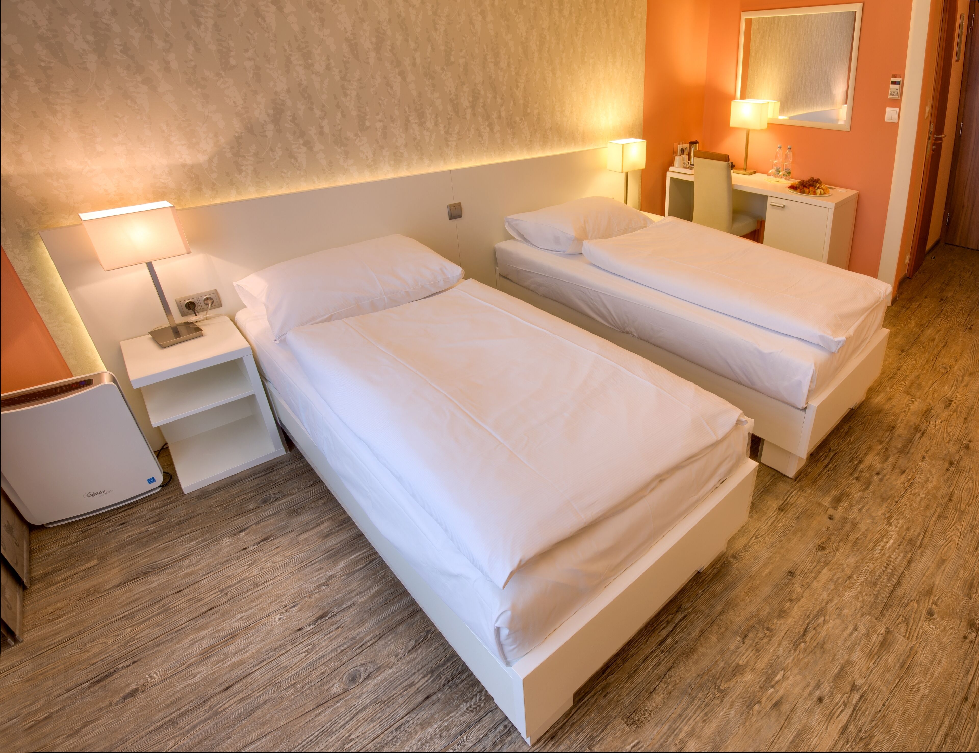 A hotel room with two single beds, a bedside table with a lamp, a small cabinet, and a wooden floor.