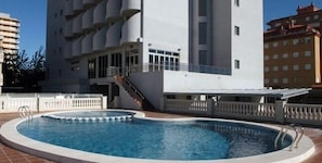 Una piscina de borde curvo, rodeada por una cerca blanca y una escalera que lleva a un edificio con balcones.