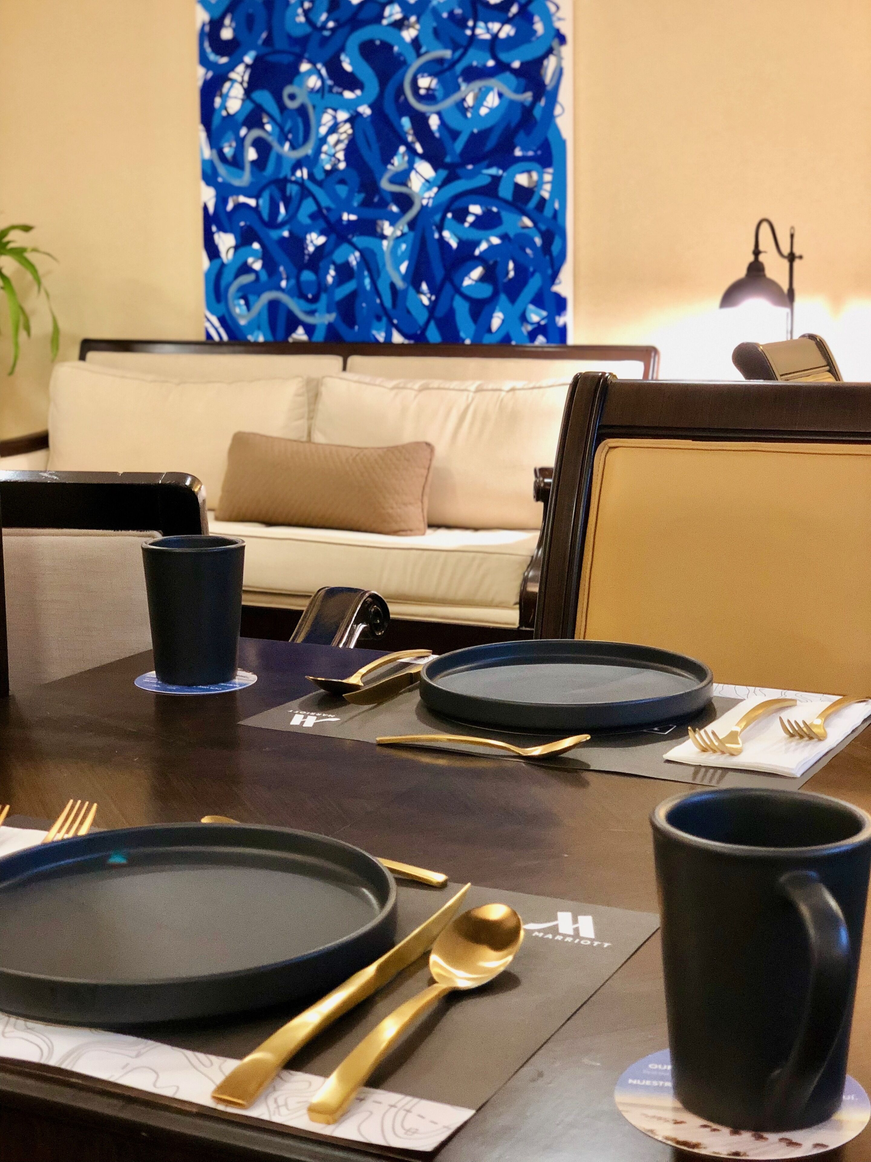 A dining table set with plates, cutlery, and a mug, with a sofa and a decorative wall in the background.