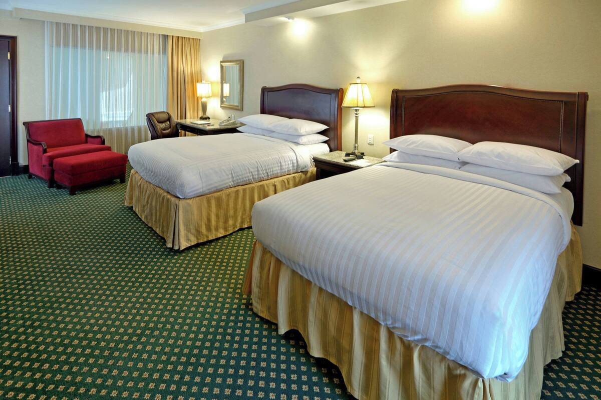 A hotel room with two beds, a red armchair, a desk with a lamp, and a large window with curtains.