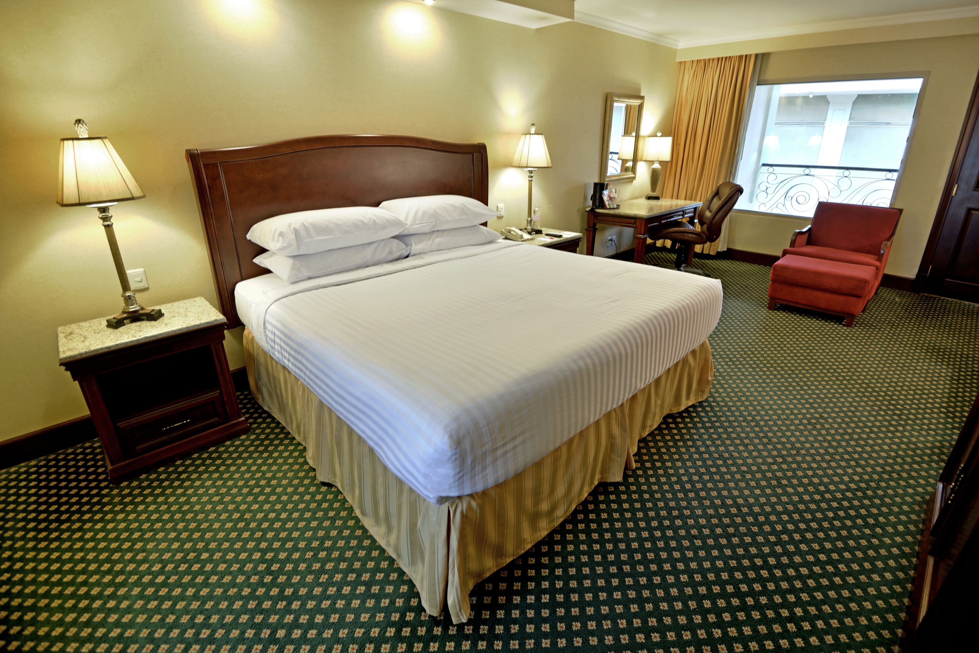 A hotel room with a large bed, two bedside tables with lamps, a desk with a chair, and a view of the outside through a window.