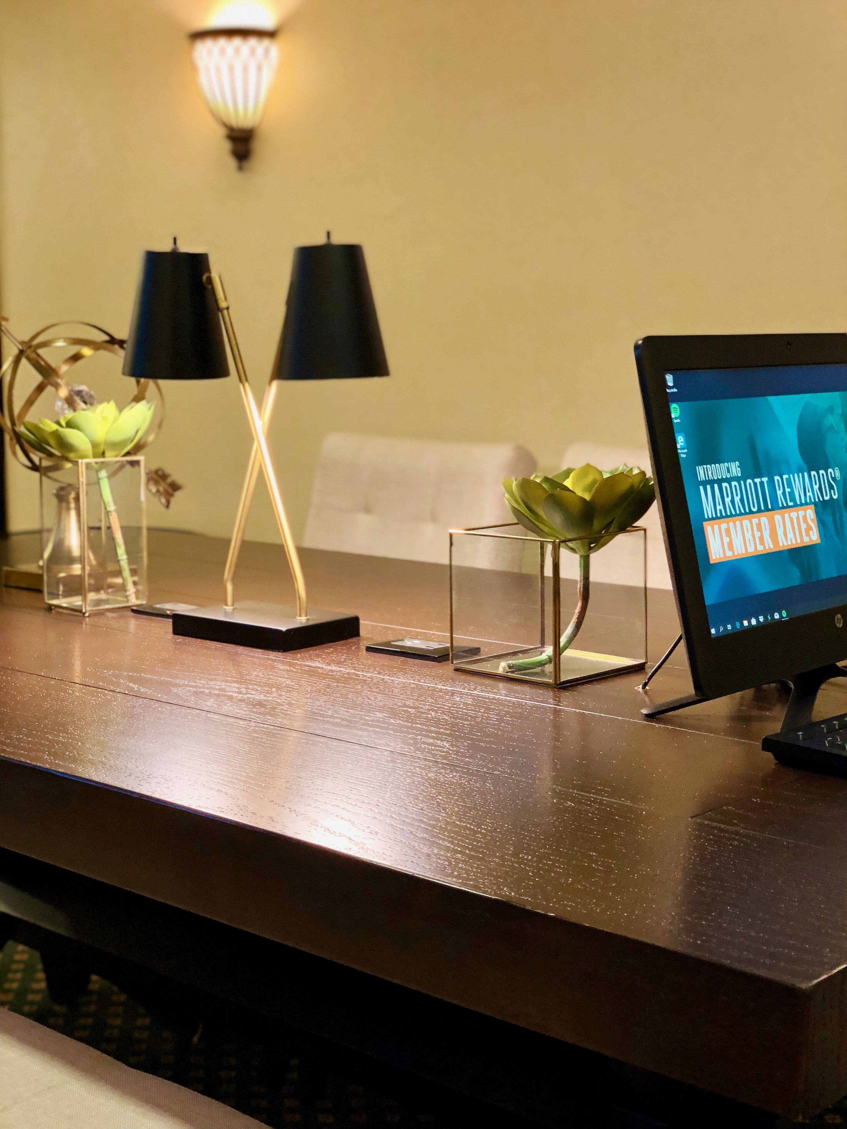 Ein Hotelrezeptionstresen mit einem Computermonitor, der 'Marriott Rewards Member Rates' anzeigt. Zwei schwarze Lampen, eine Pflanze in einer Vase und ein kleiner Tisch mit einem dekorativen Gegenstand sind ebenfalls vorhanden.