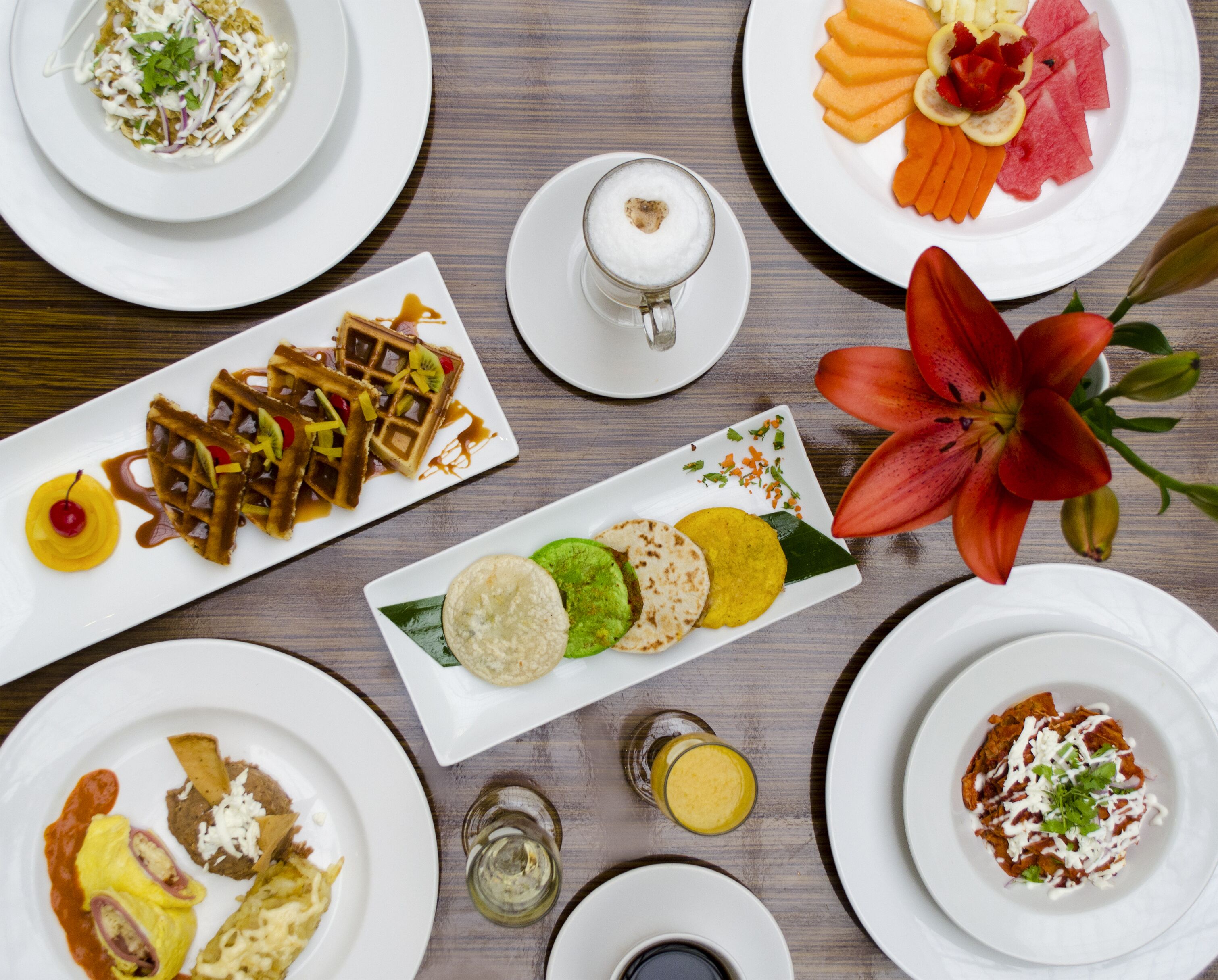 Diversos pratos sobre uma mesa de madeira, incluindo waffles, frutas e uma flor.