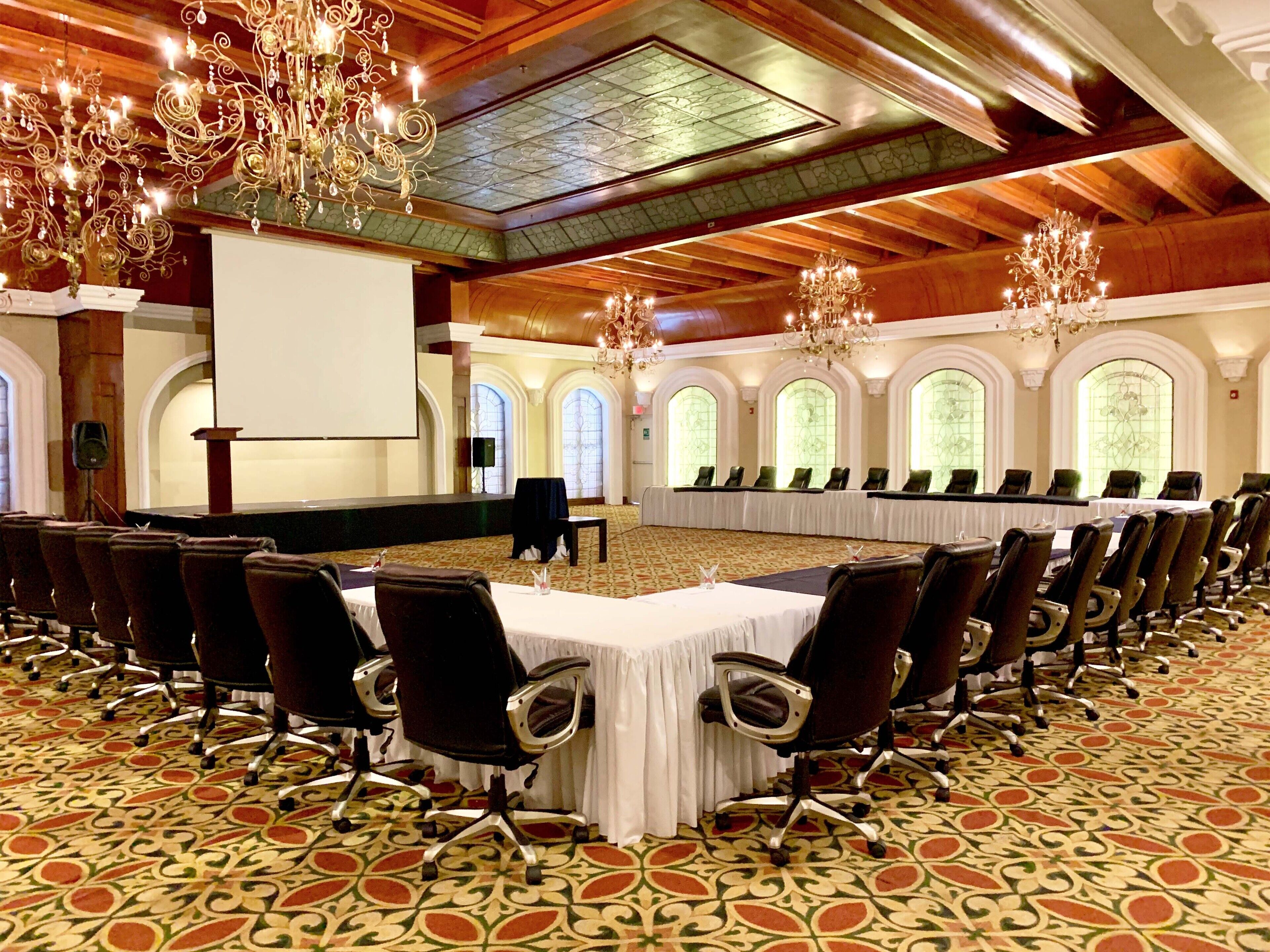 Un salón de conferencias con una larga mesa, filas de sillas, candelabros y ventanas de arco.