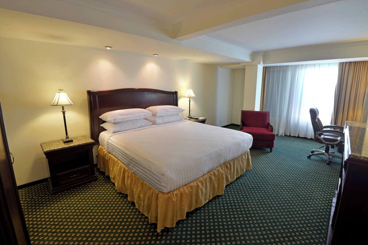 A hotel room with a large bed, two bedside lamps, a red chair, and a desk with a chair.