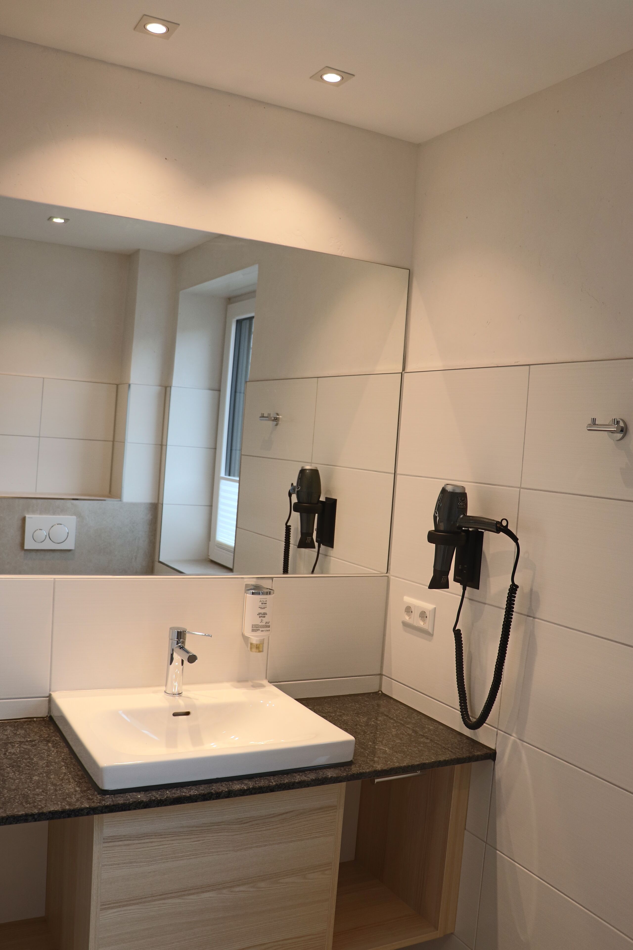 Ein modernes Badezimmer mit einem großen Spiegel, einem weißen Waschbecken und einem an der Wand befestigten Föhn.